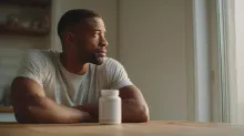 A thoughtful man sitting at a wooden table with a protein supplement bottle in front of him, looking out the window.