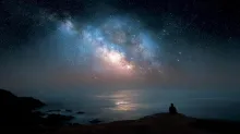 A lone man sitting on a cliff above the ocean at night, looking at the Milky Way galaxy in a vast starry sky.